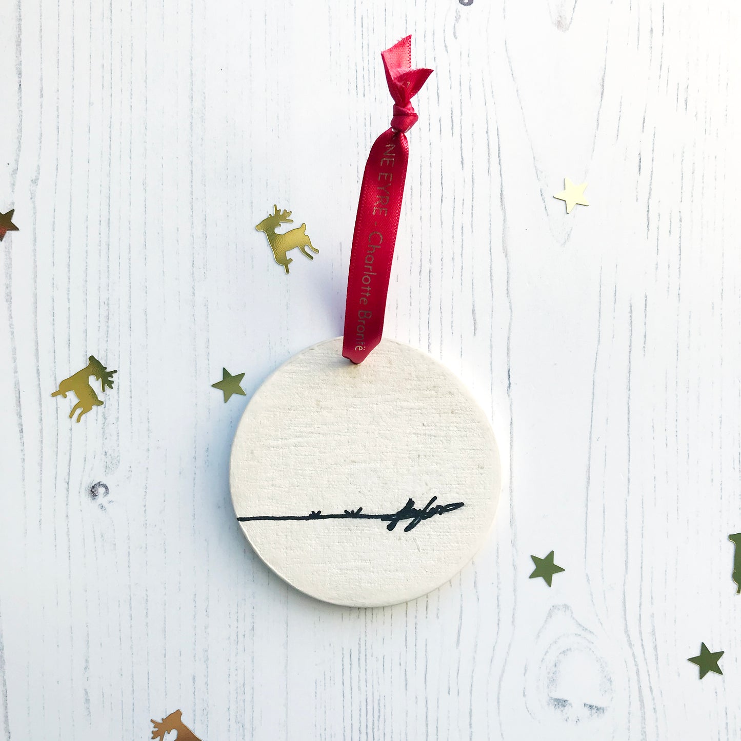 The back of a circular white ceramic hanging decoration showing a hand painted black artists signature. It hangs from a satin red ribbon with gold foil lettering that says Jane Eyre - Charlotte Brontë.