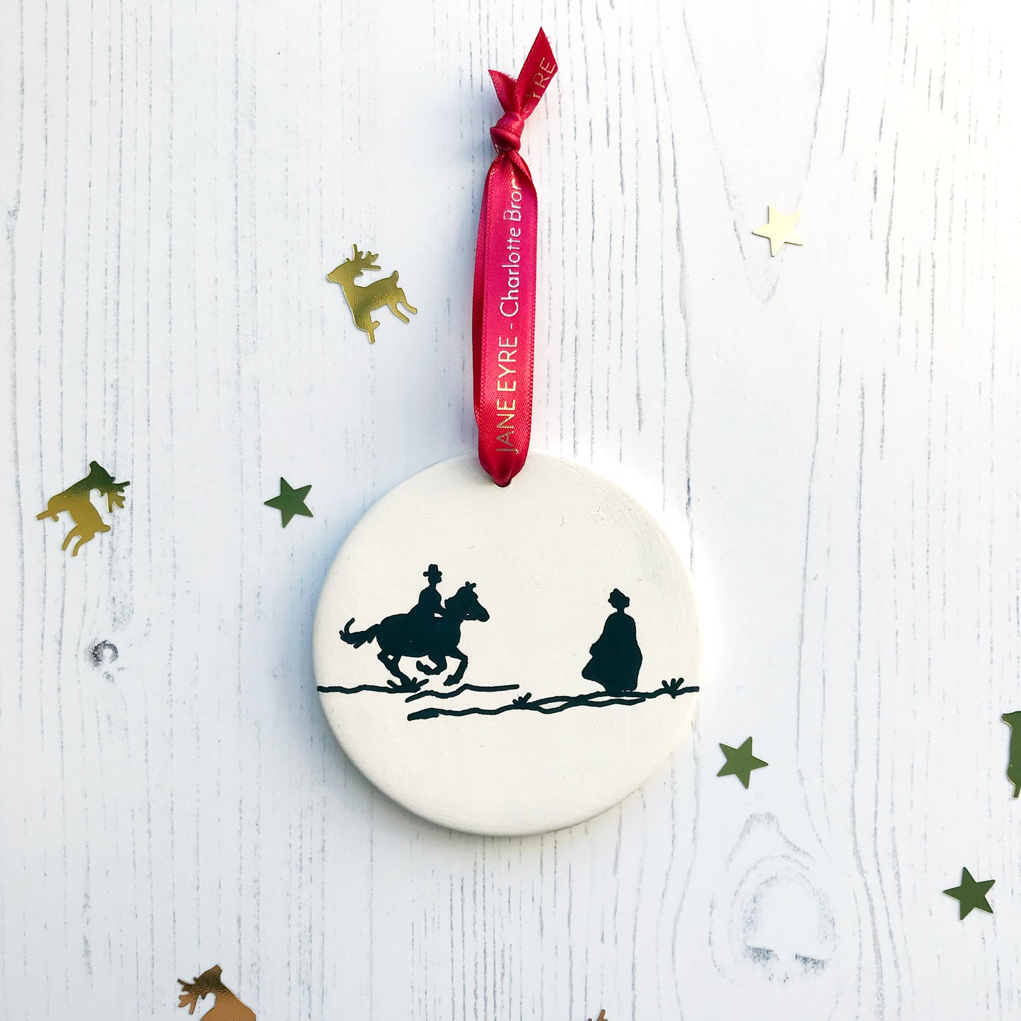 A circular white ceramic hanging decoration showing a hand painted black silhouette scene inspired by the novel Jane Eyre by Charlotte Brontë. The scene shows a silhouette scene of a man galloping on a horse towards a woman, Mr Rochester and Jane. They are in Victorian dress. It hangs from a satin red ribbon with gold foil lettering that says Jane Eyre - Charlotte Brontë.