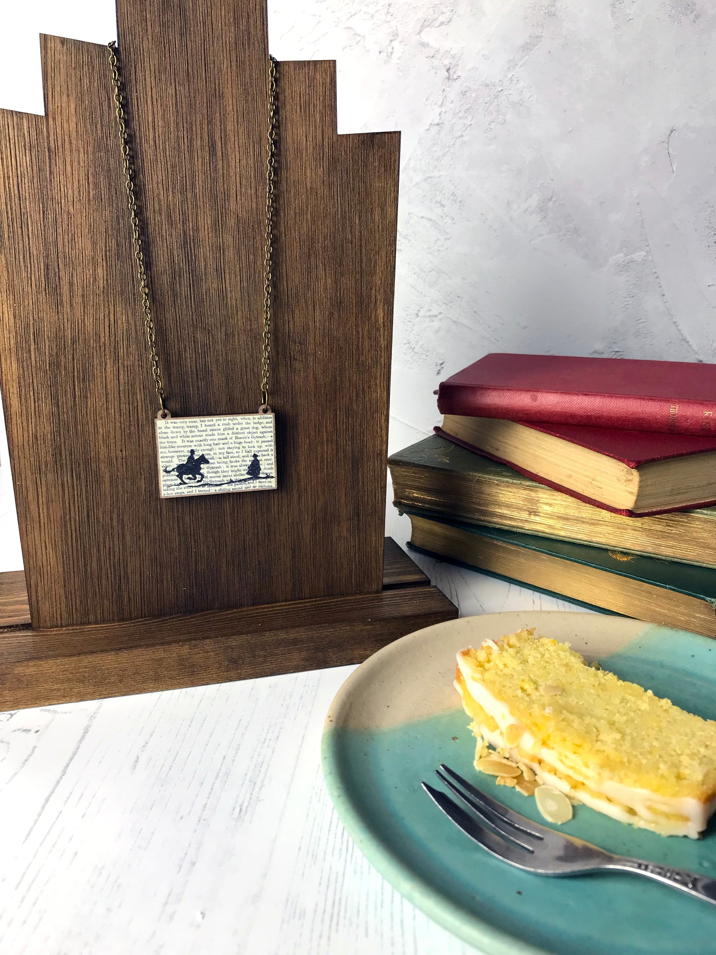 A laser cut wooden necklace by loadofolbobbins showing a silhouette scene inspired by the novel Jane Eyre by Charlotte Brontë. The scene shows a silhouette scene of a man galloping on a horse towards a woman, Mr Rochester and Jane. They are in Victorian dress. The scene is printed against original text from the novel. The necklace is hung over a wooden display form.