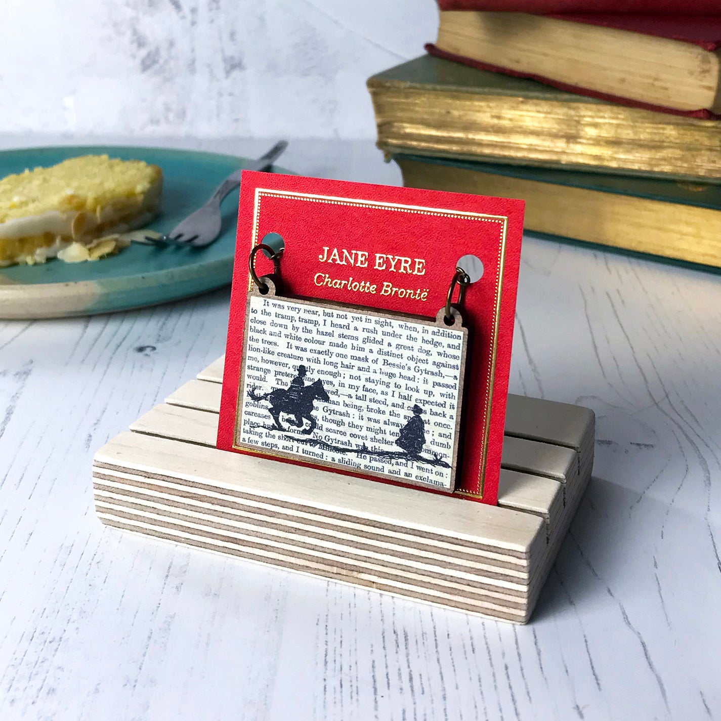 A laser cut wooden necklace by loadofolbobbins showing a silhouette scene inspired by the novel Jane Eyre by Charlotte Brontë. The scene shows a silhouette scene of a man galloping on a horse towards a woman, Mr Rochester and Jane. They are in Victorian dress. The scene is printed against original text from the novel. The necklace is mounted on a red backing card with gold foil lettering detail and made to look like the front of a book.