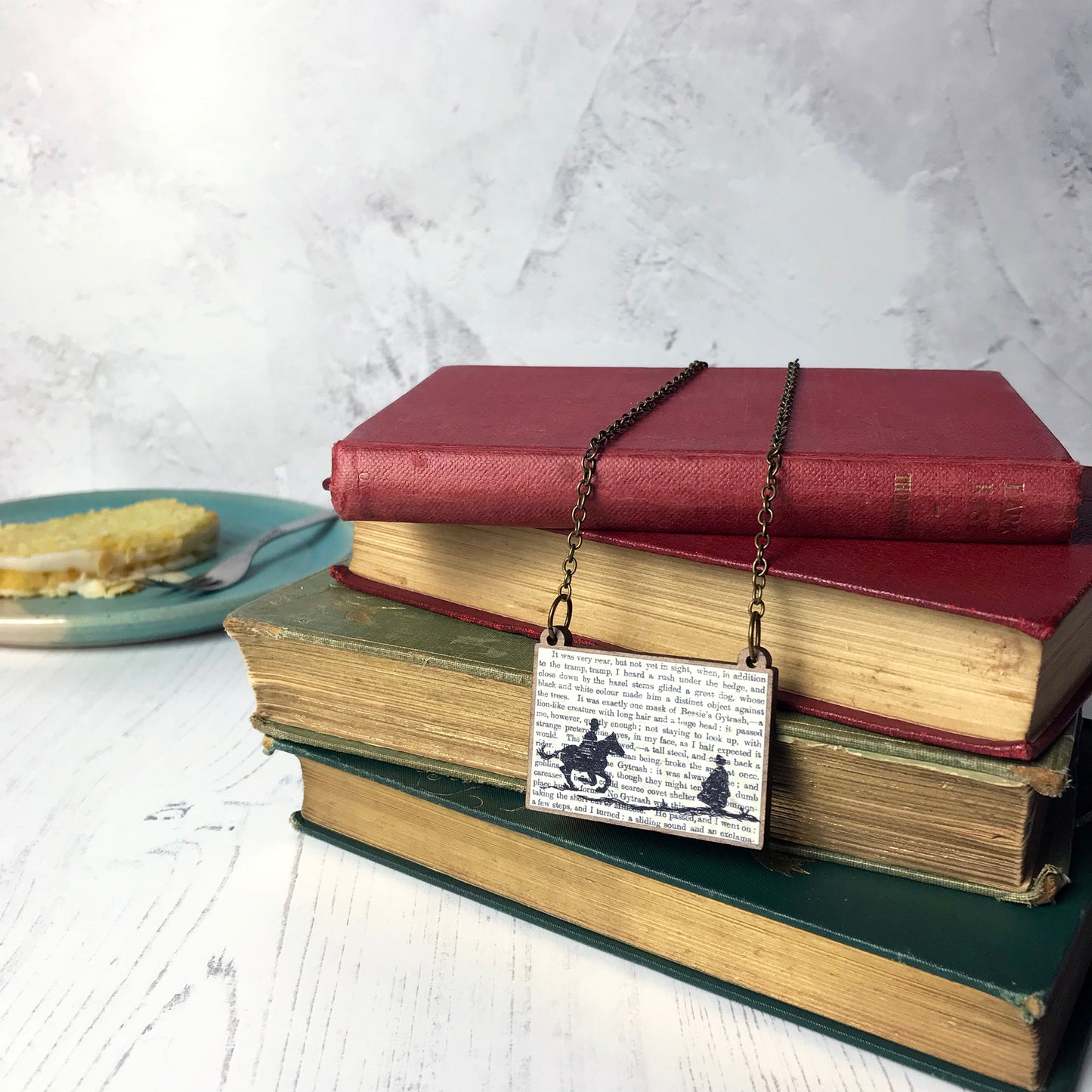 A laser cut wooden necklace by loadofolbobbins showing a silhouette scene inspired by the novel Jane Eyre by Charlotte Brontë. The scene shows a silhouette scene of a man galloping on a horse towards a woman, Mr Rochester and Jane. They are in Victorian dress. The scene is printed against original text from the novel. The necklace is draped over and dangling on a pile of old books.