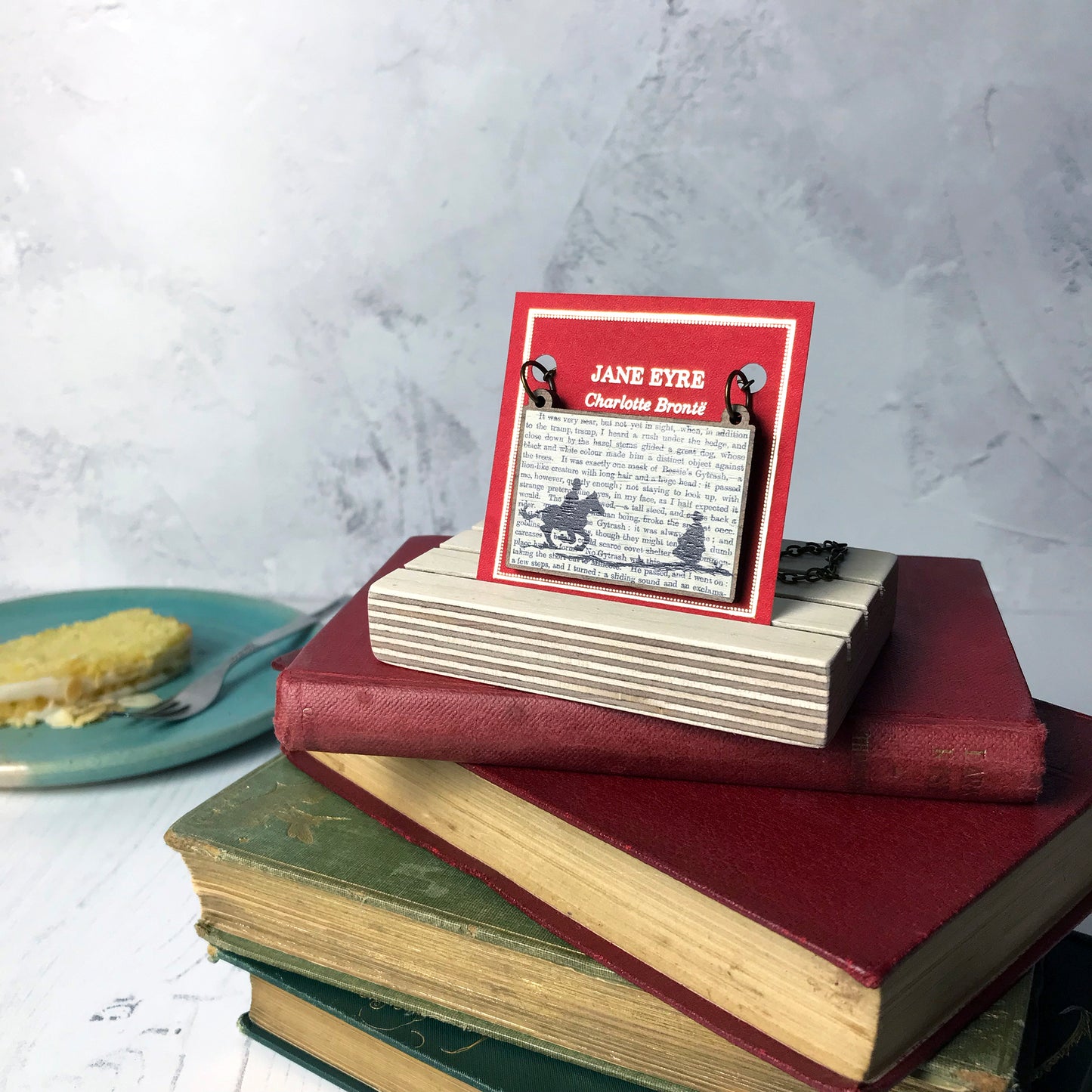 A laser cut wooden necklace by loadofolbobbins showing a silhouette scene inspired by the novel Jane Eyre by Charlotte Brontë. The scene shows a silhouette scene of a man galloping on a horse towards a woman, Mr Rochester and Jane. They are in Victorian dress. The scene is printed against original text from the novel. The necklace is mounted on a red backing card with gold foil lettering detail and made to look like the front of a book and is standing on a pile of old books.