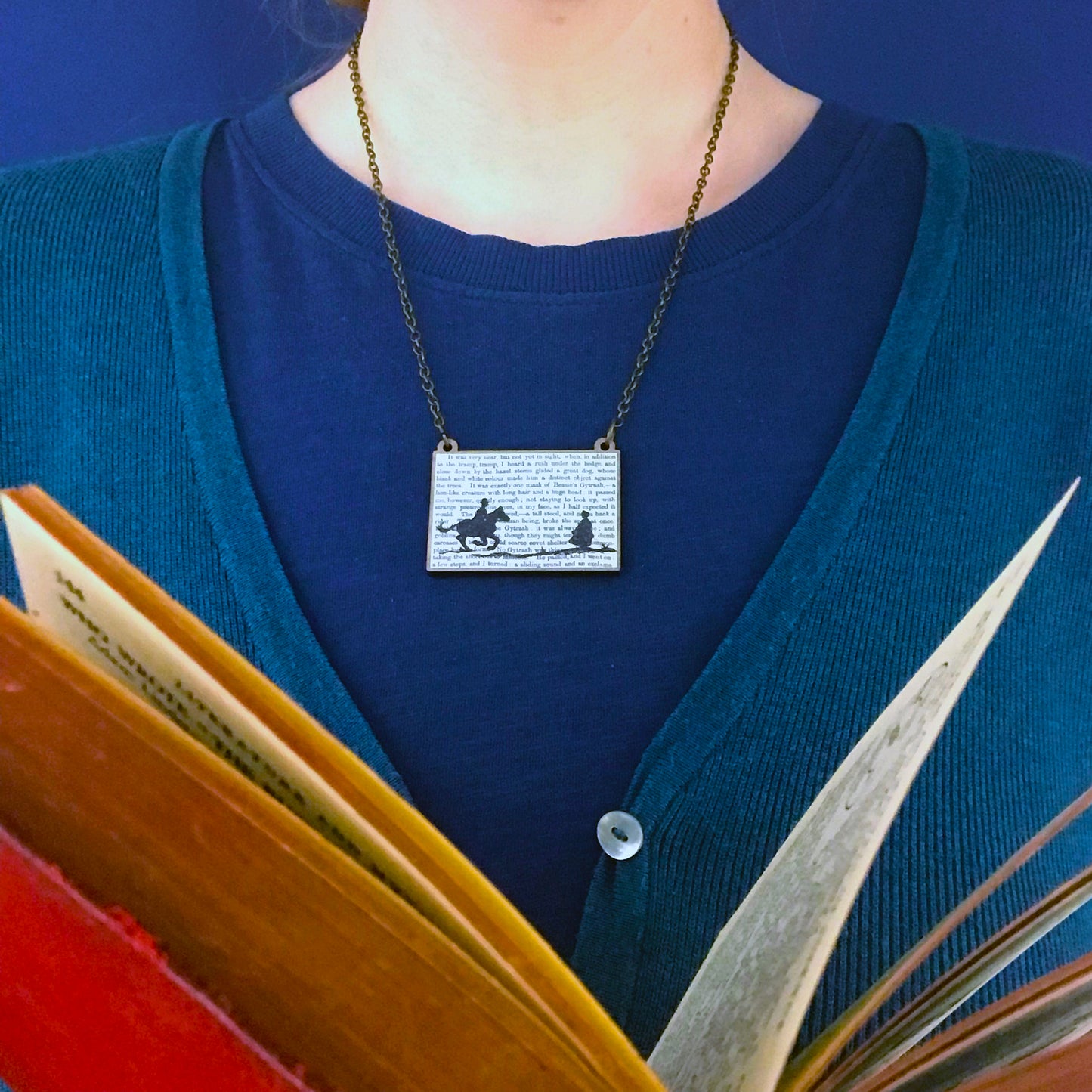 A laser cut wooden necklace by loadofolbobbins showing a silhouette scene inspired by the novel Jane Eyre by Charlotte Brontë. The scene shows a silhouette scene of a man galloping on a horse towards a woman, Mr Rochester and Jane. They are in Victorian dress. The scene is printed against original text from the novel. The necklace is being worn against a blue top.