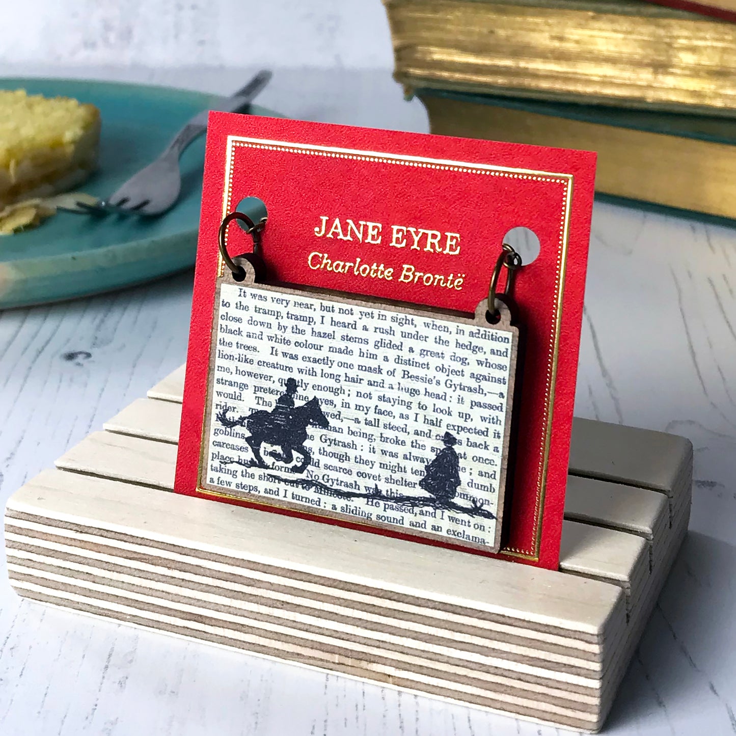 A laser cut wooden necklace by loadofolbobbins showing a silhouette scene inspired by the novel Jane Eyre by Charlotte Brontë. The scene shows a silhouette scene of a man galloping on a horse towards a woman, Mr Rochester and Jane. They are in Victorian dress. The scene is printed against original text from the novel. The necklace is mounted on a red backing card with gold foil lettering detail and made to look like the front of a book.