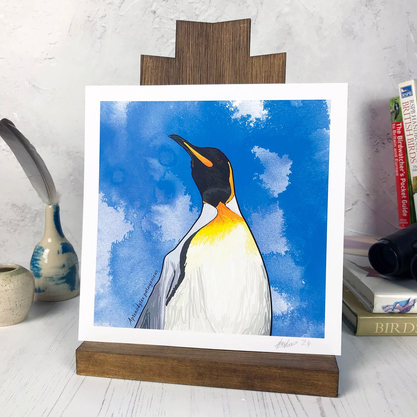 A photo of a medium sized 21 cm square art print made from an original digital illustration by Loadofolbobbins. It is of a king penguin with energetic pen line marks. The illustration is a close up of the shoulders and head and is set against a vibrant blue ink wash background. It has a very regal like pose and the viewer is almost looking up at the penguin. It has a cream belly which leads to vibrant yellow orange colouring, black head and beak and grey back. There is a 2.5 cm white border for framing.
