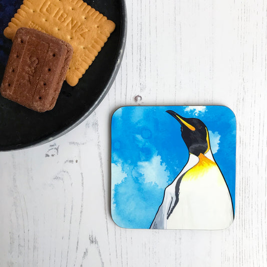 A photo of a of round edged square coaster which has an original illustration by Loadofolbobbins showing a king penguin with energetic pen line marks. The illustration is a close up of the shoulders and head and is set against a vibrant blue ink wash background. It has a very regal like pose and the viewer is almost looking up at the penguin. It has a cream belly which leads to vibrant yellow orange colouring, black head and beak and grey back.