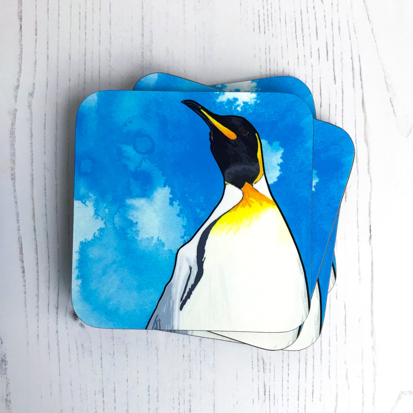A photo of a pile of round edged square coasters which have an original illustration by Loadofolbobbins showing a king penguin with energetic pen line marks. The illustration is a close up of the shoulders and head and is set against a vibrant blue ink wash background. It has a very regal like pose and the viewer is almost looking up at the penguin. It has a cream belly which leads to vibrant yellow orange colouring, black head and beak and grey back.