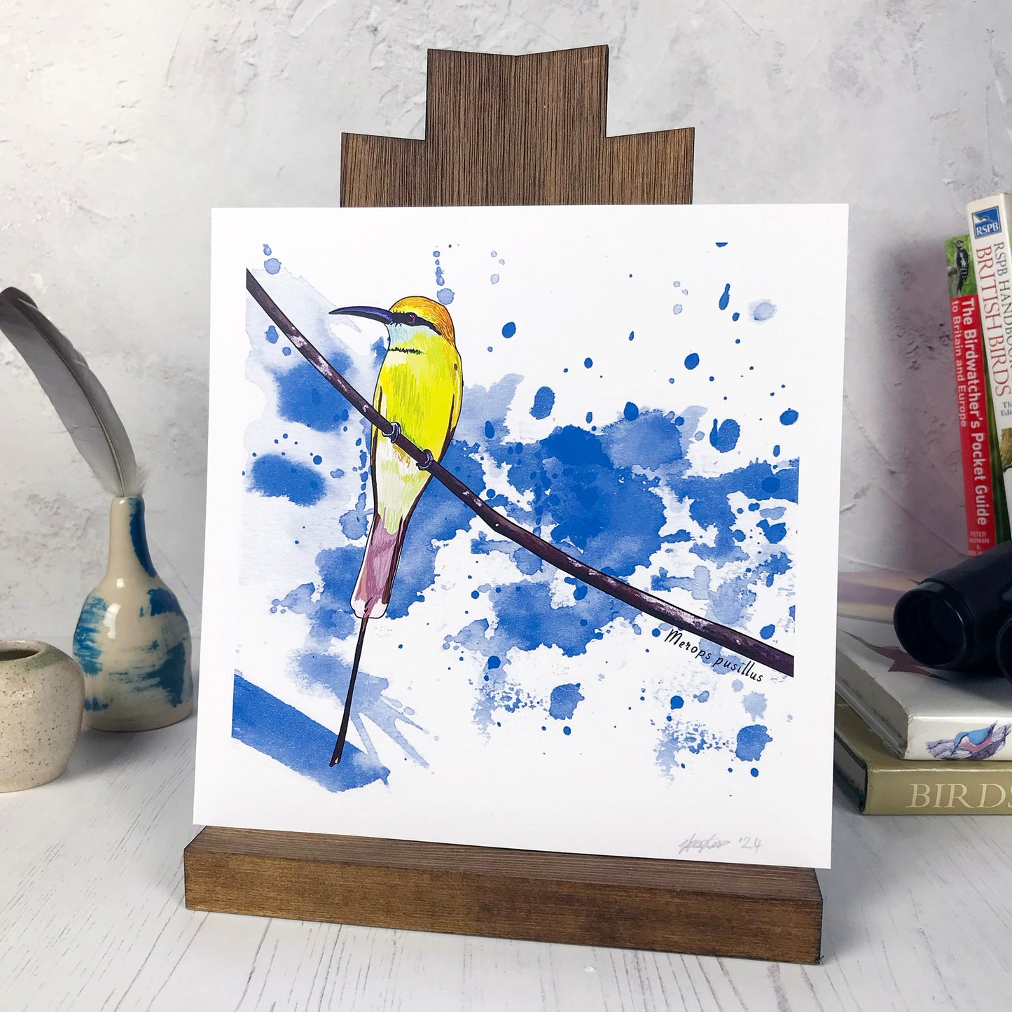 A photo of a medium sized 21 cm square art print made from an original illustration by Loadofolbobbins. It is of a little bee eater bird perched on a fine curved branch. They have vibrant green upper feathers and yellow head. there's a black band of feathers like a zorro mask. It has a long pointed curved grey beak. The background of the image is made of rich bright blue inky splash marks. There is a 2.5 cm white border for framing.