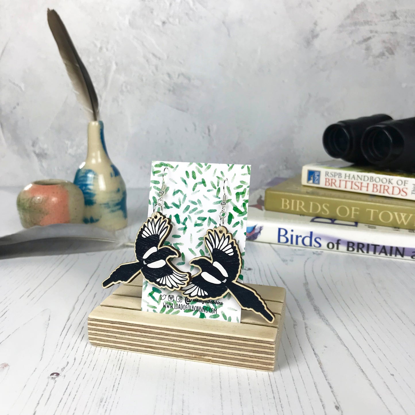 A pair of laser cut wooden dangle earrings showing an illustration of a flying magpie bird by loadofolbobbins. The earrings are mounted on a backing card with a repeat abstract watercolour leaf pattern. They are standing in a wooden stand and in the background you can see a pile of bird watching books and a pair of binoculars.
