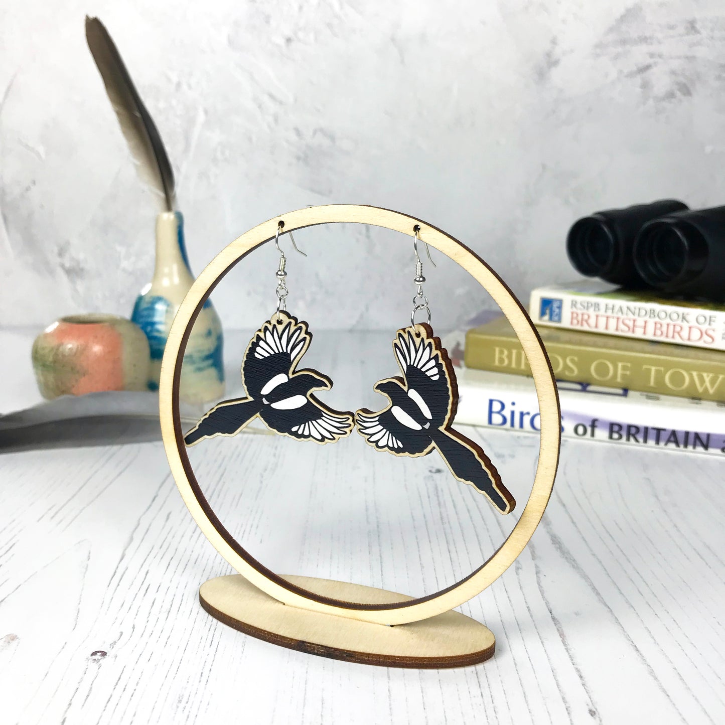 A pair of laser cut wooden dangle earrings showing an illustration of a flying magpie bird by loadofolbobbins. The earrings are hanging from a wooden circular ring display.