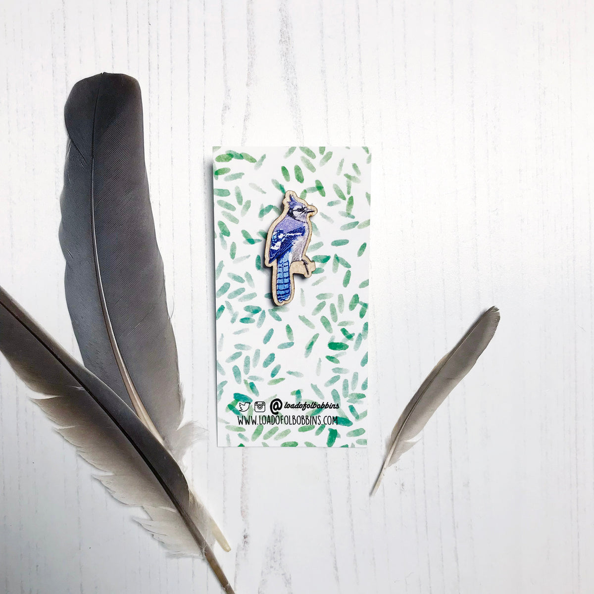 A 33mm mini wooden lapel pin of a Blue jay bird perched on a branch and mounted on an illustrated backing card. The pin is made from an original illustration and is detailed and expressive in style. The bird is a wonderful array of blues with a small crest on it's head. The backing card has a repeat green watercolour effect pattern that looks like leaves.