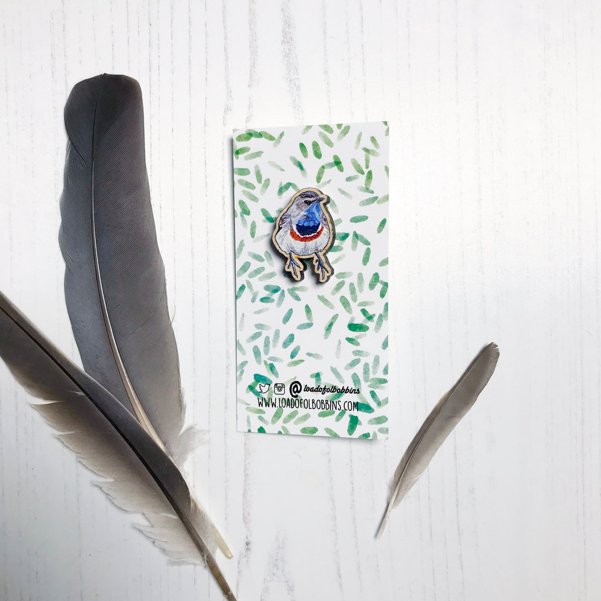 A 25mm mini wooden lapel pin of a Bluethroat bird mounted on an illustrated backing card. The pin is made from an original illustration and is detailed and expressive in style. The bird is rather robin like in appearance but with a bright blue throat. The backing card has a repeat green watercolour effect pattern that looks like leaves.