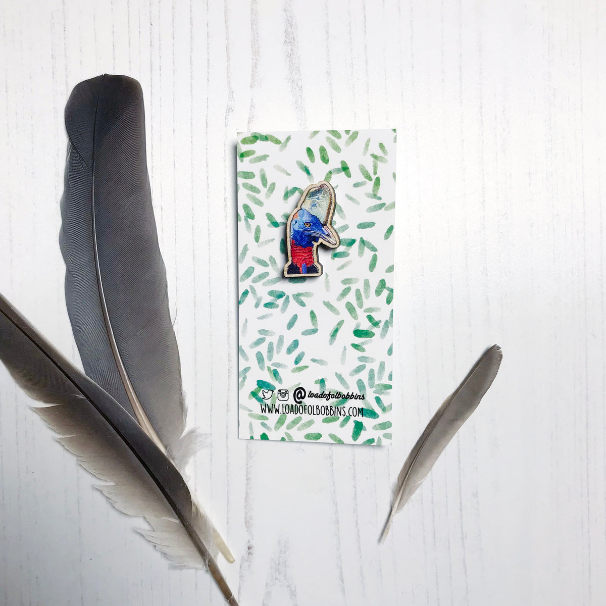 A 25mm mini wooden lapel pin of the head of a Cassowary bird mounted on an illustrated backing card. The pin is made from an original illustration and is detailed and expressive in style. The bird is rather dinosaur like in appearance with a bright blue head and red neck with blue green casque on it's head. The backing card has a repeat green watercolour effect pattern that looks like leaves.