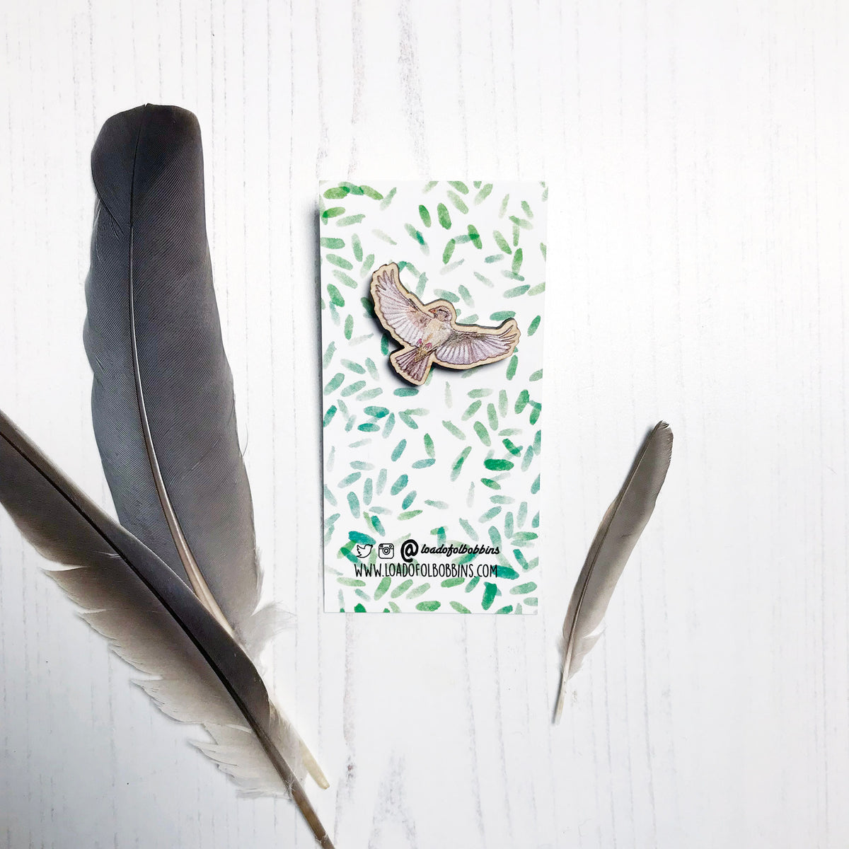 A 30mm mini wooden lapel pin of a flying female sparrow mounted on an illustrated backing card. The pin is made from an original illustration and is detailed and expressive in style. The bird is a gorgeous array of brown hues and has its wings spread wide beside it. The backing card has a repeat green watercolour effect pattern that looks like leaves.