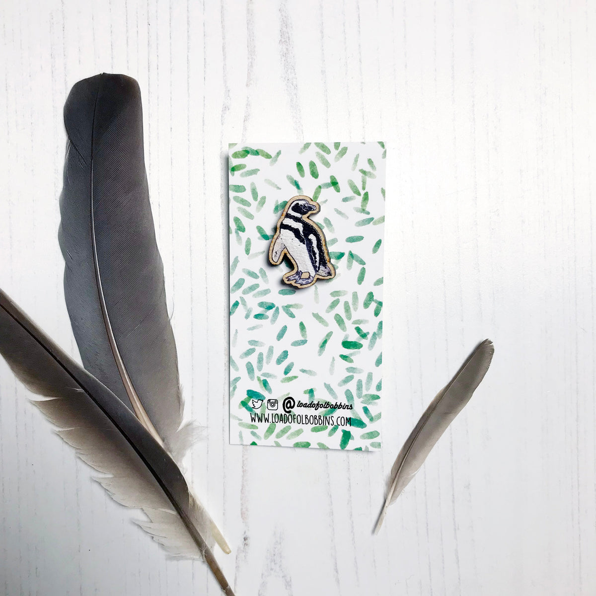 A 27mm mini wooden lapel pin of a magellanic penguin mounted on an illustrated backing card. The pin is made from an original illustration and is detailed and expressive in style. The bird has black and white markings. The backing card has a repeat green watercolour effect pattern that looks like leaves.