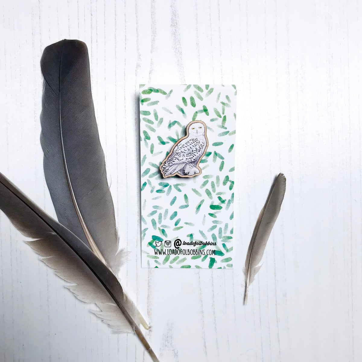 A 25mm mini wooden lapel pin of a snowy owl mounted on an illustrated backing card. The pin is made from an original illustration and is detailed and expressive in style. The bird snow white with brown flecked markings. The backing card has a repeat green watercolour effect pattern that looks like leaves.