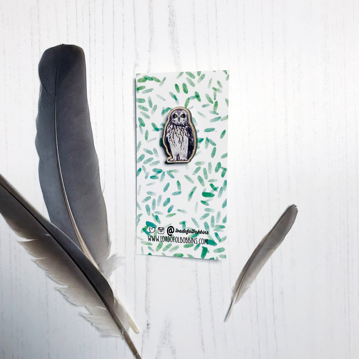 A 25mm mini wooden lapel pin of a short-eared owl mounted on an illustrated backing card. The pin is made from an original illustration and is detailed and expressive in style. The bird dark brown wings and cream front with brown flecked markings. The backing card has a repeat green watercolour effect pattern that looks like leaves.