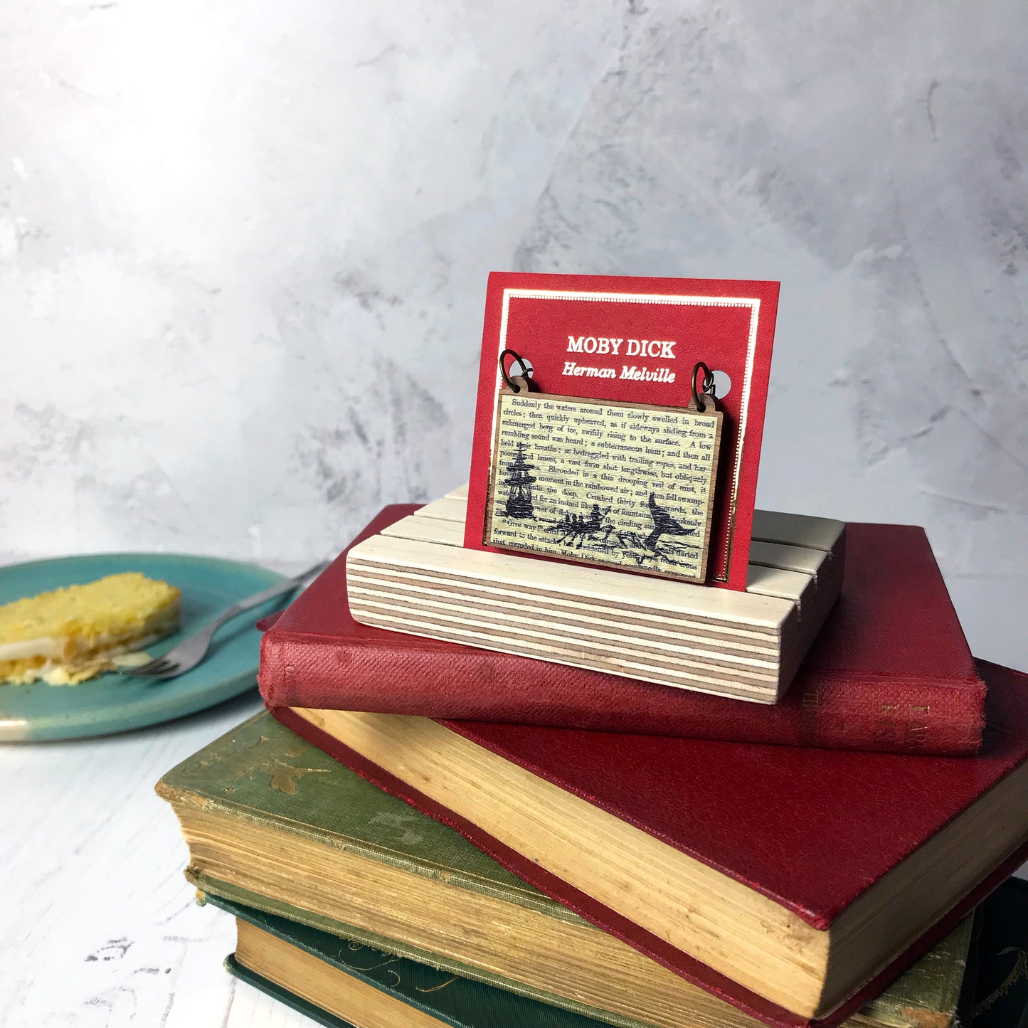 A laser cut wooden necklace showing a silhouette scene of a sailing ship and a rowing boat with men holding spears in it chasing after a large whale tail as it dives below the sea. It is inspired by the novel Moby Dick by Herman Melville and is printed with the text from the novel behind it. The necklace is mounted on a red backing card with gold foil lettering detail and made to look like the front of a book and put on a pile of books.