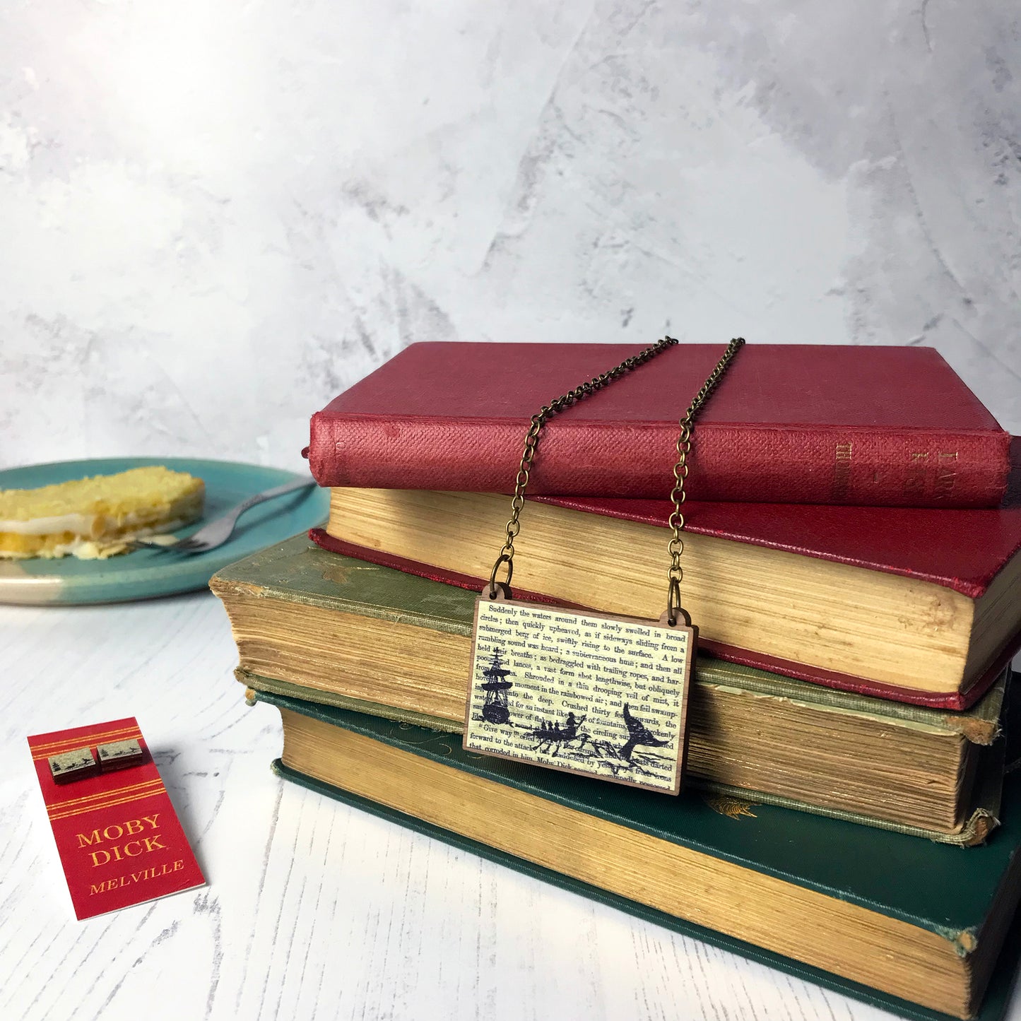 Laser cut wooden necklace and stud earrings showing a silhouette scene of a sailing ship and a rowing boat with men holding spears in it chasing after a large whale tail as it dives below the sea. It is inspired by the novel Moby Dick by Herman Melville and is printed with the text from the novel behind it. The necklace is draped over a book, the earrings are mounted on a red backing card with gold lettering detail and made to look like the spine of a book.