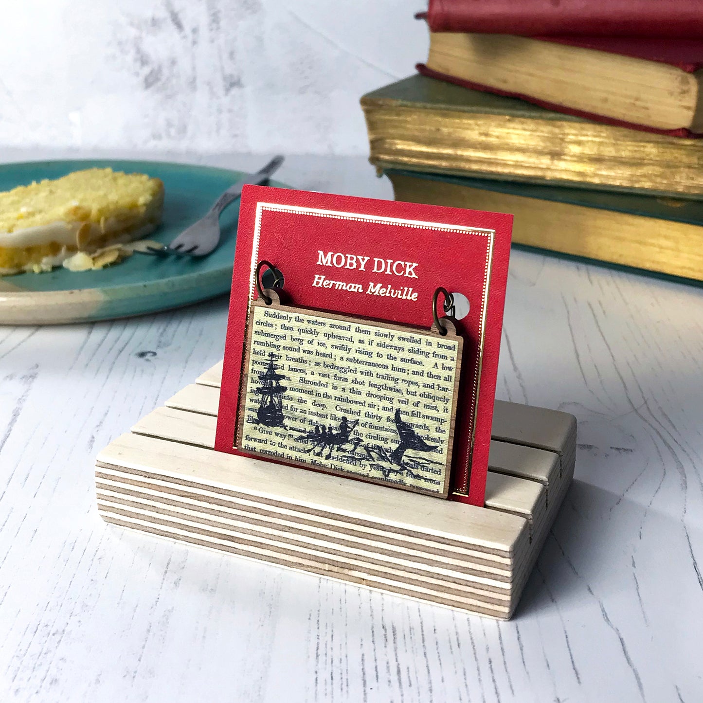 A laser cut wooden necklace showing a silhouette scene of a sailing ship and a rowing boat with men holding spears in it chasing after a large whale tail as it dives below the sea. It is inspired by the novel Moby Dick by Herman Melville and is printed with the text from the novel behind it. The necklace is mounted on a red backing card with gold foil lettering detail and made to look like the front of a book.