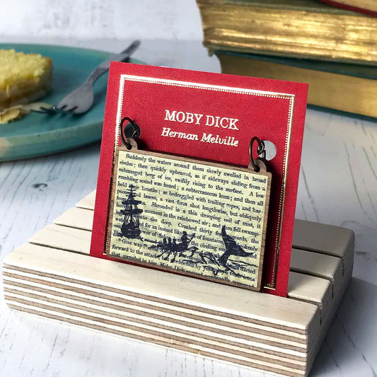 A laser cut wooden necklace showing a silhouette scene of a sailing ship and a rowing boat with men holding spears in it chasing after a large whale tail as it dives below the sea. It is inspired by the novel Moby Dick by Herman Melville and is printed with the text from the novel behind it. The necklace is mounted on a red backing card with gold foil lettering detail and made to look like the front of a book.