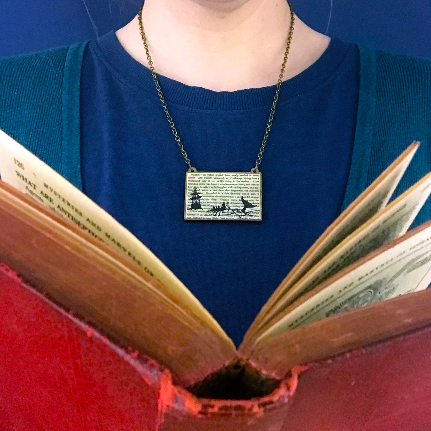 A laser cut wooden necklace showing a silhouette scene of a sailing ship and a rowing boat with men holding spears in it chasing after a large whale tail as it dives below the sea. It is inspired by the novel Moby Dick by Herman Melville and is printed with the text from the novel behind it. The necklace is being worn against a blue top.