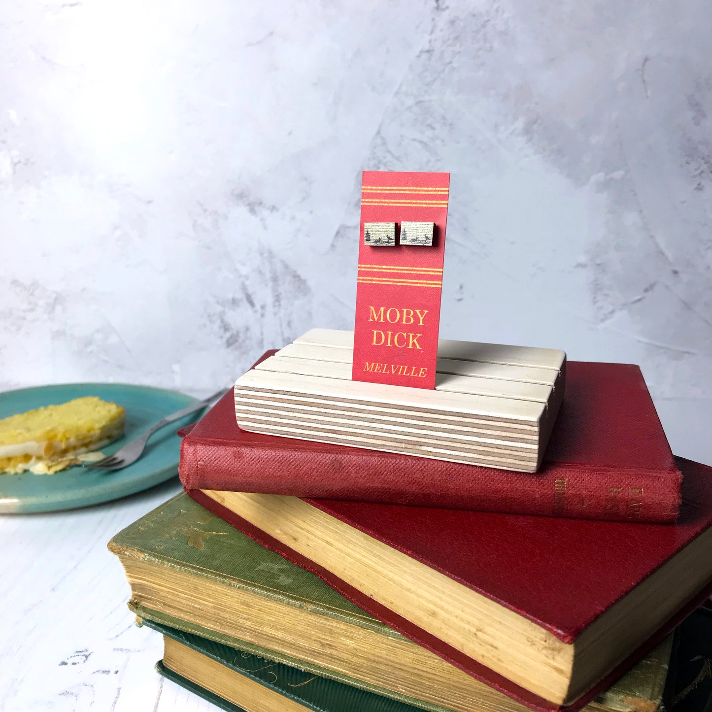 Laser cut wooden stud earrings by loadofolbobbins showing a silhouette scene inspired by the book Moby Dick by Herman Melville. It shows a silhouette scene of a sailing ship and a rowing boat with men holding spears in it chasing after a large whale tail as it dives below the sea. The scene is printed against original text from the novel. The earrings are mounted on a red backing card with gold lettering detail and made to look like the spine of a book.