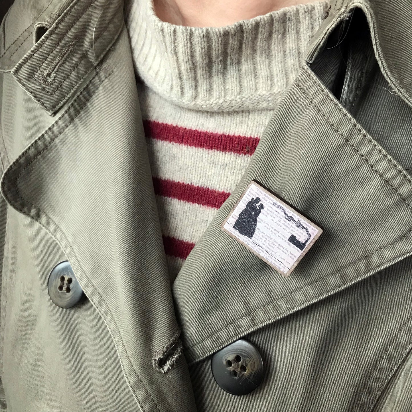 A laser cut wooden brooch showing a silhouette scene of a couple in victorian dress embracing and a factory in the background with a billowing chimney. It is inspired by the novel North and South by Elizabeth Gaskell and is printed with the text from the novel behind it. The brooch is being worn on the lapel of a jacket.