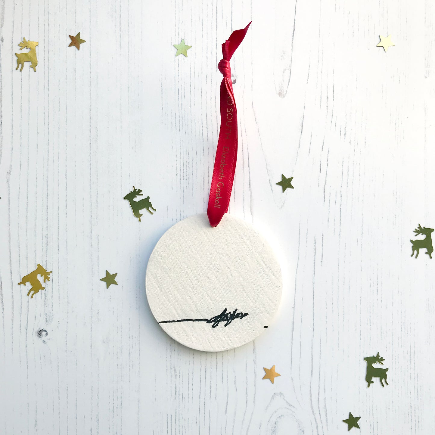 The back of a circular white ceramic hanging decoration showing a hand painted black artists signature. It hangs from a satin red ribbon with gold foil lettering that says North and South - Elizabeth Gaskell.