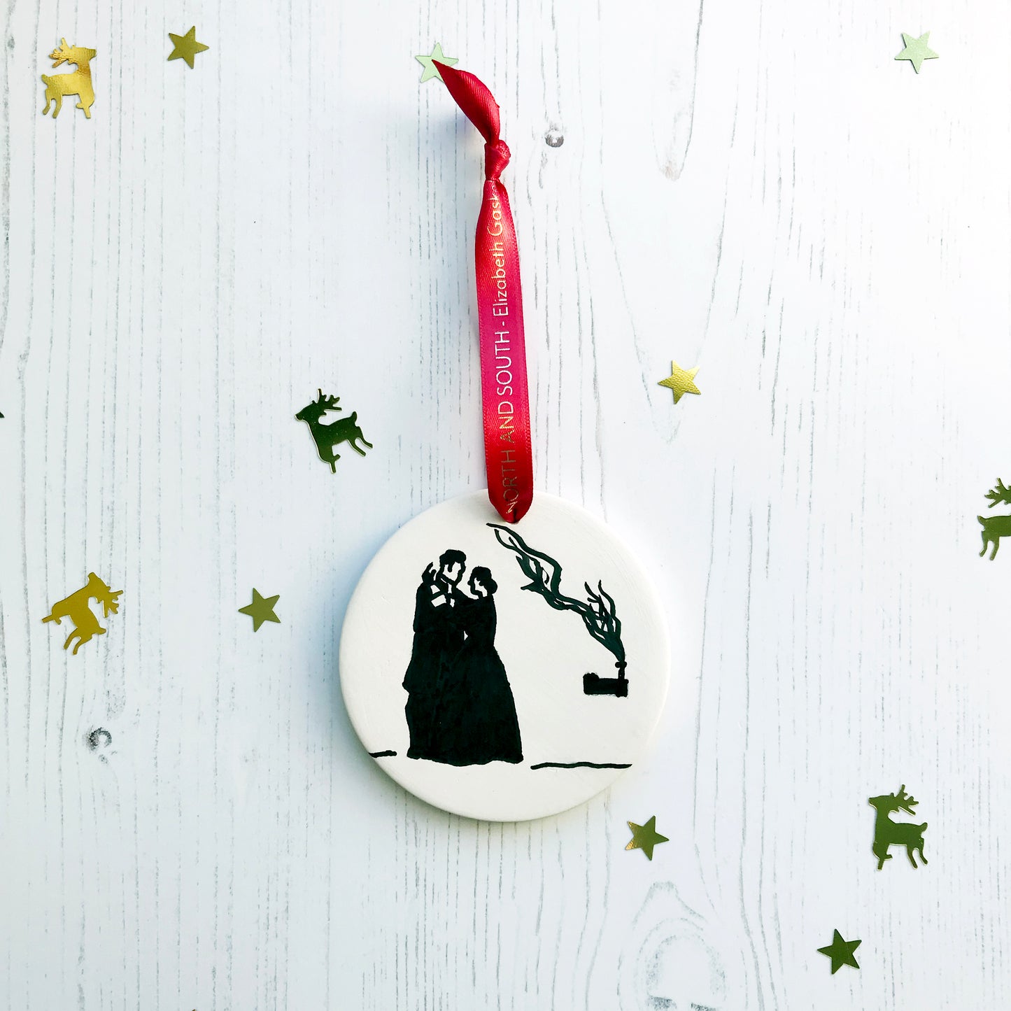 A circular white ceramic hanging decoration showing a hand painted black silhouette scene inspired the novel North and South by Elizabeth Gaskell. The scene shows Margaret Hale and Mr Thornton embracing with a industrial building with a smoking chimney of in the distance to the right of them. It hangs from a satin red ribbon with gold foil lettering that says North and South - Elizabeth Gaskell.