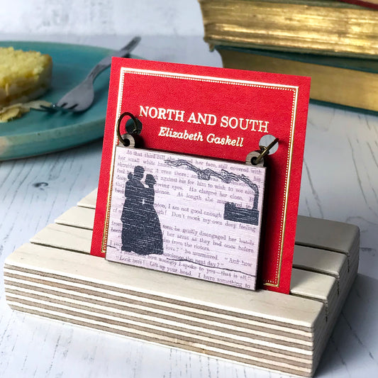 A laser cut wooden necklace showing a silhouette scene a couple in Victorian dress embracing and a factory in the background with a billowing chimney. It is inspired by the novel North and South by Elizabeth Gaskell and is printed with the text from the novel behind it. The necklace is mounted on a red backing card with gold foil lettering detail and made to look like the front of a book.