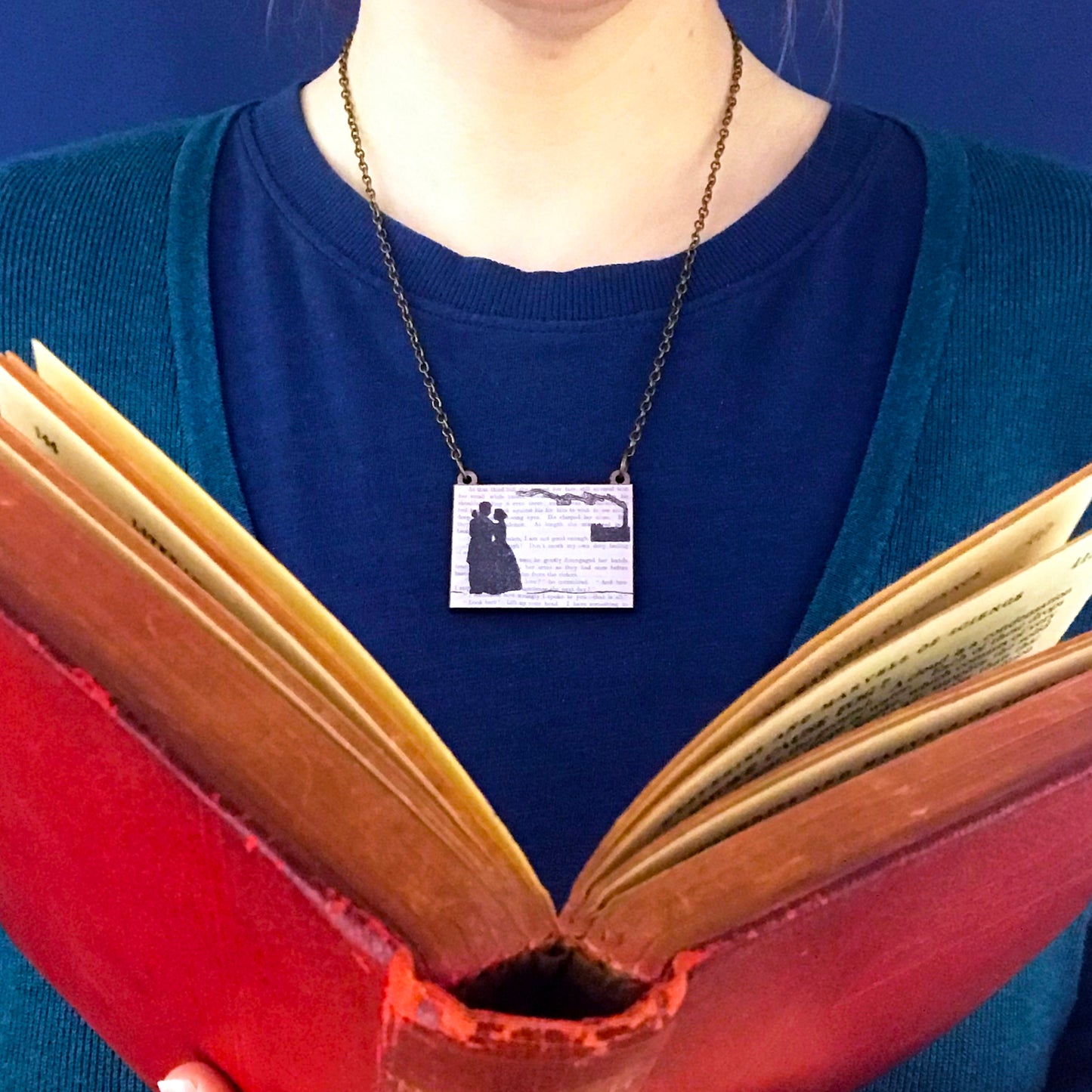 A laser cut wooden necklace showing a silhouette scene of a couple in victorian dress embracing and a factory in the background with a billowing chimney. It is inspired by the novel North and South by Elizabeth Gaskell and is printed with the text from the novel behind it. The necklace is being worn against a blue top.