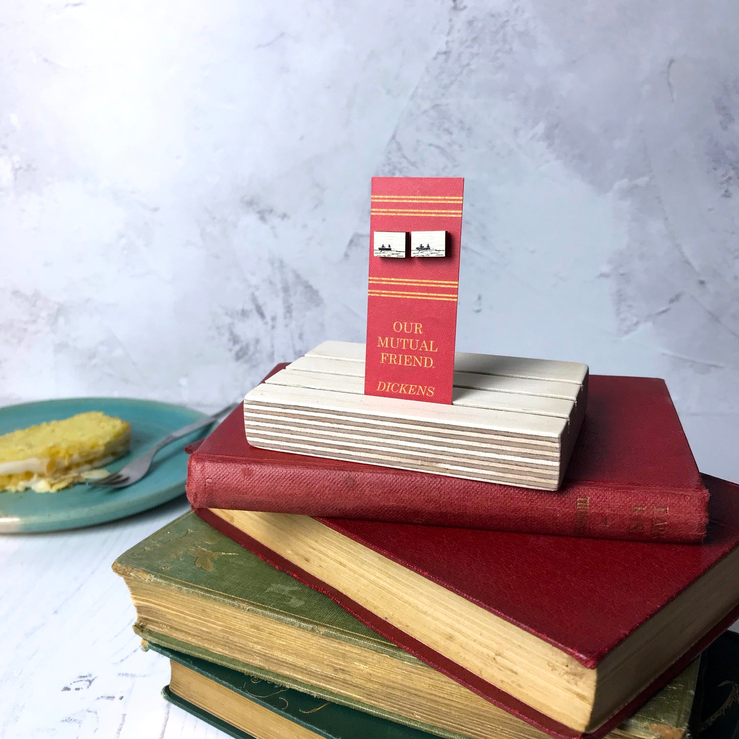 Laser cut wooden stud earrings by loadofolbobbins showing showing a silhouette scene inspired by the novel Our Mutual Friend by Charles Dickens. The scene shows a man and woman rowing in the Thames with a body dragged behind it, they are Lizzie Hexham and her father. The scene is printed against original text from the novel. The earrings are mounted on a red backing card with gold lettering detail and made to look like the spine of a book.