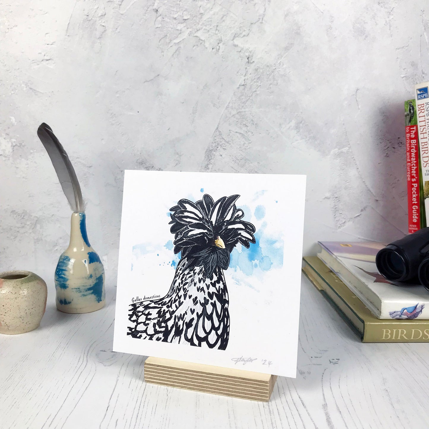 A photo of a small sized 14 cm square art print made from an original illustration by Loadofolbobbins. It is of a polish chicken. It is a close neck up illustration showing the incredible feathered head that is an explosion of black and white feathers with it’s beak just peaking out from underneath them. The background is an light blue inky splash. There is a 2.5 cm white border for framing.