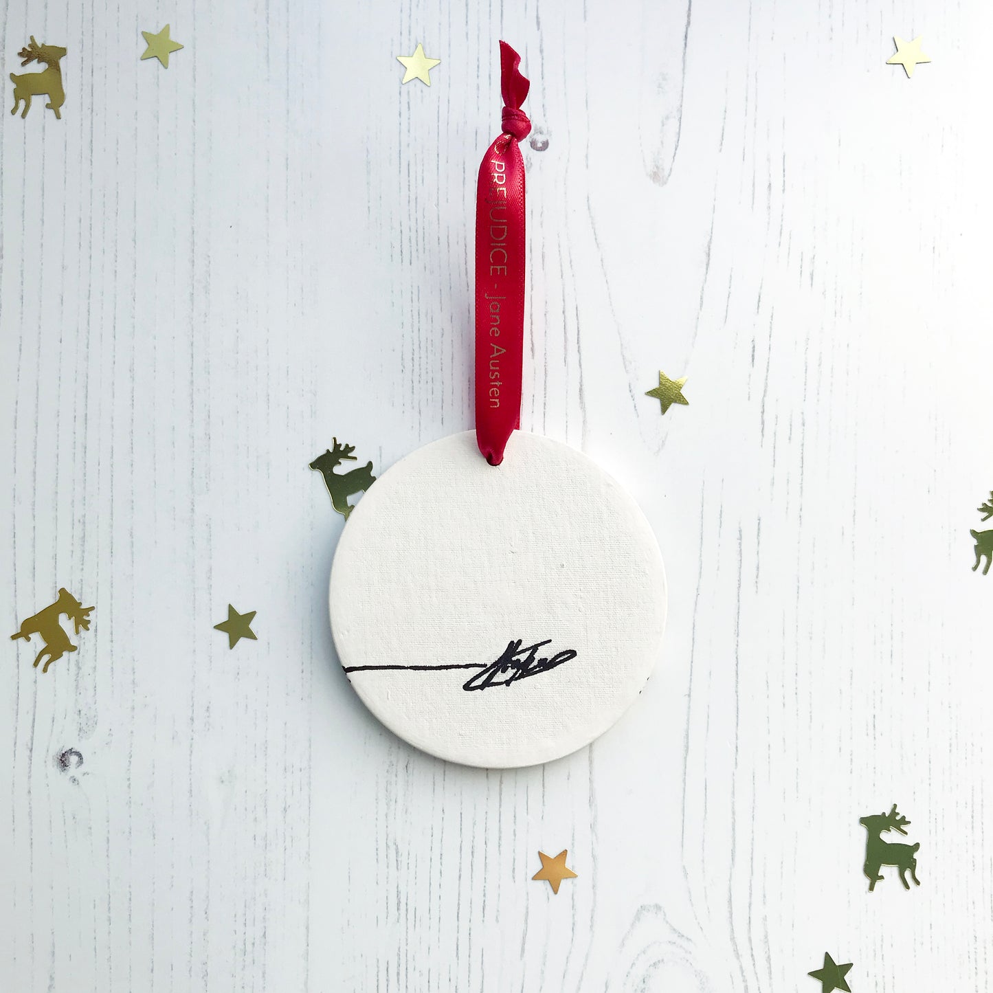 The back of a circular white ceramic hanging decoration showing a hand painted black artists signature. It hangs from a satin red ribbon with gold foil lettering that says Pride and Prejudice - Jane Austen.