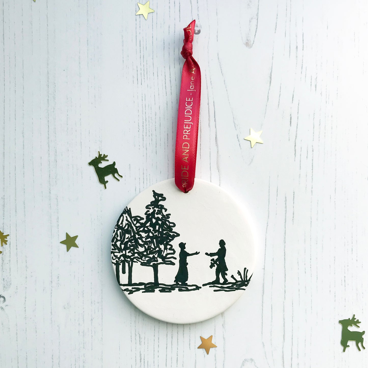 A circular white ceramic hanging decoration showing a hand painted black silhouette scene inspired by the novel Pride and Prejudice by Jane Austen. The scene shows a silhouette scene of a man and woman in regency costume. The woman is walking out of a wood towards the man who is holding out a letter to her, they are Elizabeth Bennet and Mr Darcy. It hangs from a satin red ribbon with gold foil lettering that says Pride and Prejudice - Jane Austen.