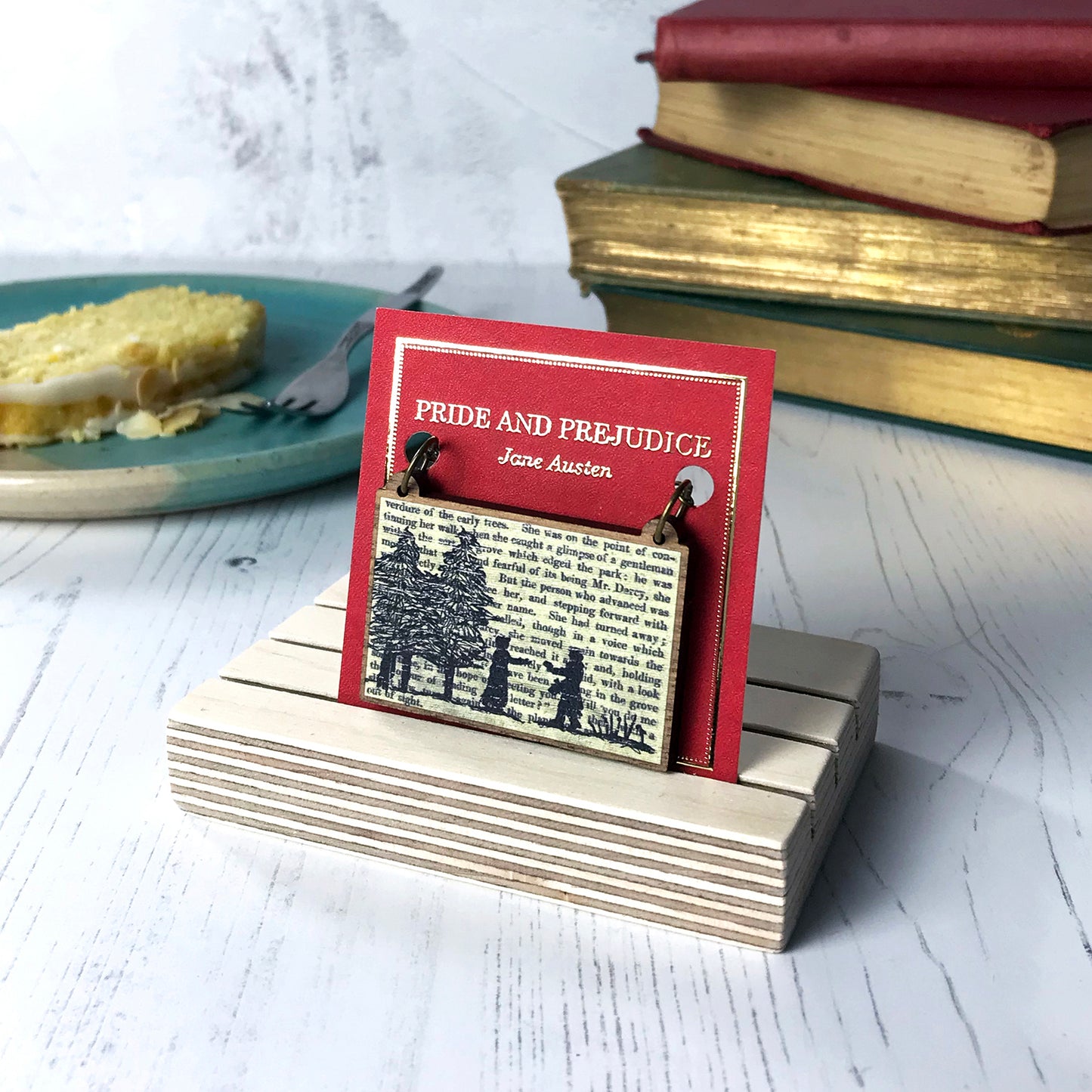 A laser cut wooden necklace by loadofolbobbins showing a silhouette scene inspired by the novel Pride and Prejudice by Jane Austen. The scene shows a silhouette scene of a man and woman in regency costume. The woman is walking out of a wood towards the man who is holding out a letter to her, they are Elizabeth Bennet and Mr Darcy. The scene is printed against original text from the novel. The necklace is mounted on a red backing card with gold foil lettering detail and made to look like the front of a book.