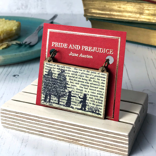 A laser cut wooden necklace by loadofolbobbins showing a silhouette scene inspired by the novel Pride and Prejudice by Jane Austen. The scene shows a silhouette scene of a man and woman in regency costume. The woman is walking out of a wood towards the man who is holding out a letter to her, they are Elizabeth Bennet and Mr Darcy. The scene is printed against original text from the novel. The necklace is mounted on a red backing card with gold foil lettering detail and made to look like the front of a book.