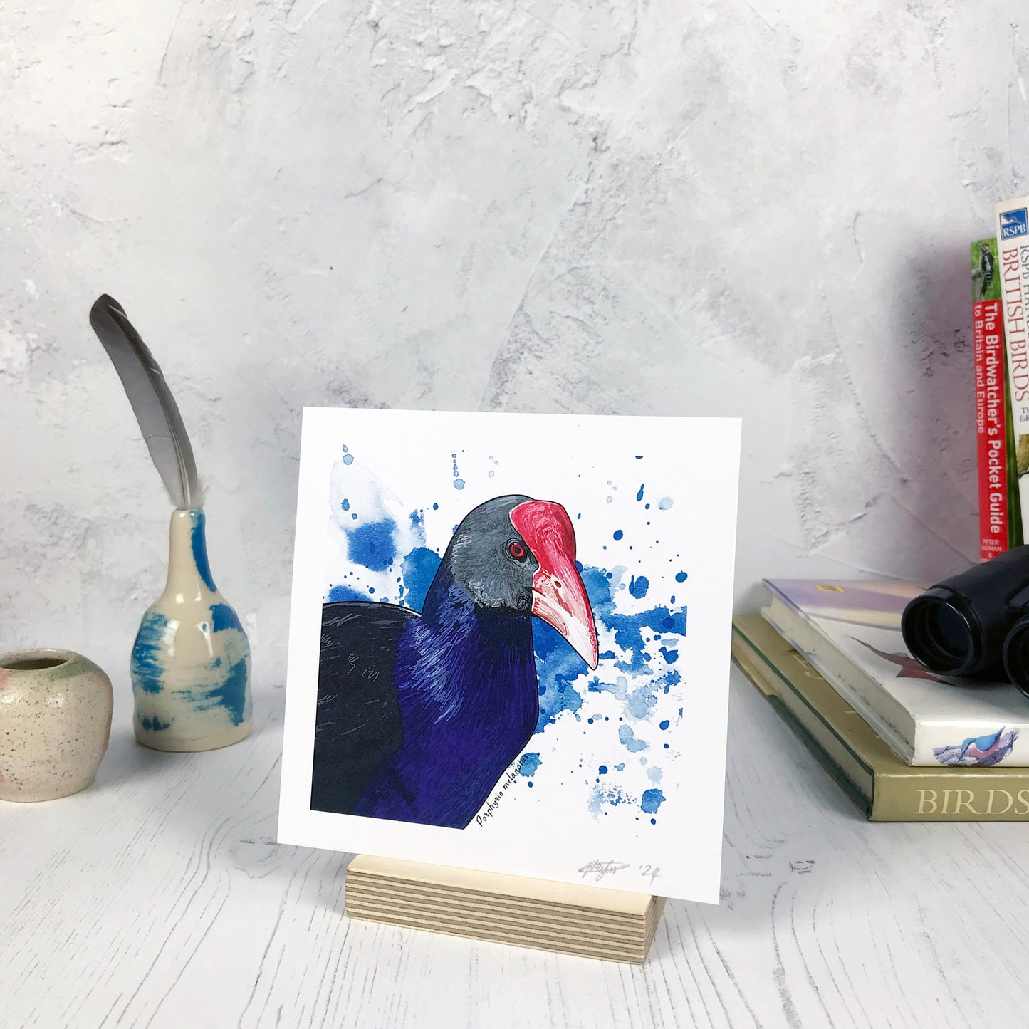 A photo of a small sized 14 cm square art print made from an original digital illustration by Loadofolbobbins. It is of a a pūkeko bird focusing on its face and upper body. It has striking deep blue feathers on it’s chest and neck with black feathered back and face. It’s red beak extends up to its forehead fanning out in shape and is bright red. The background is a dark blue inky splash. There is a 2.5 cm white border for framing.