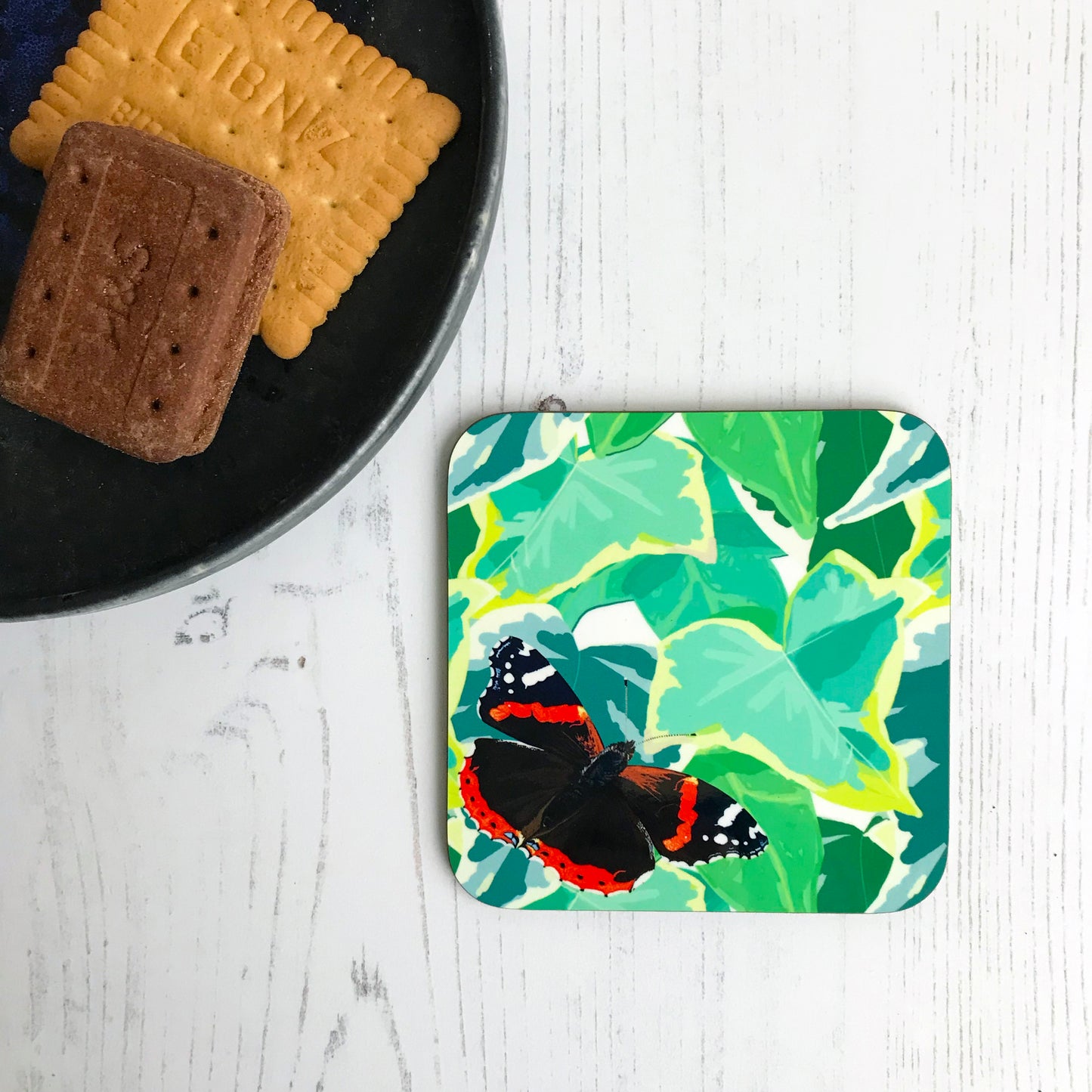 A photo of a round edged square coaster which has an original illustration by Loadofolbobbins. It shows a red admiral butterfly that's landed on a mass of vibrant green ivy. It's wings are spread out beside it showing it's beautiful markings, the red stripes midway in the upper wings and at the bottom of it's lower wings. The tips of the upper wings are black with white marking and dots. The butterfly is in the bottom left corner of the coaster with the ivy covering the rest of it.