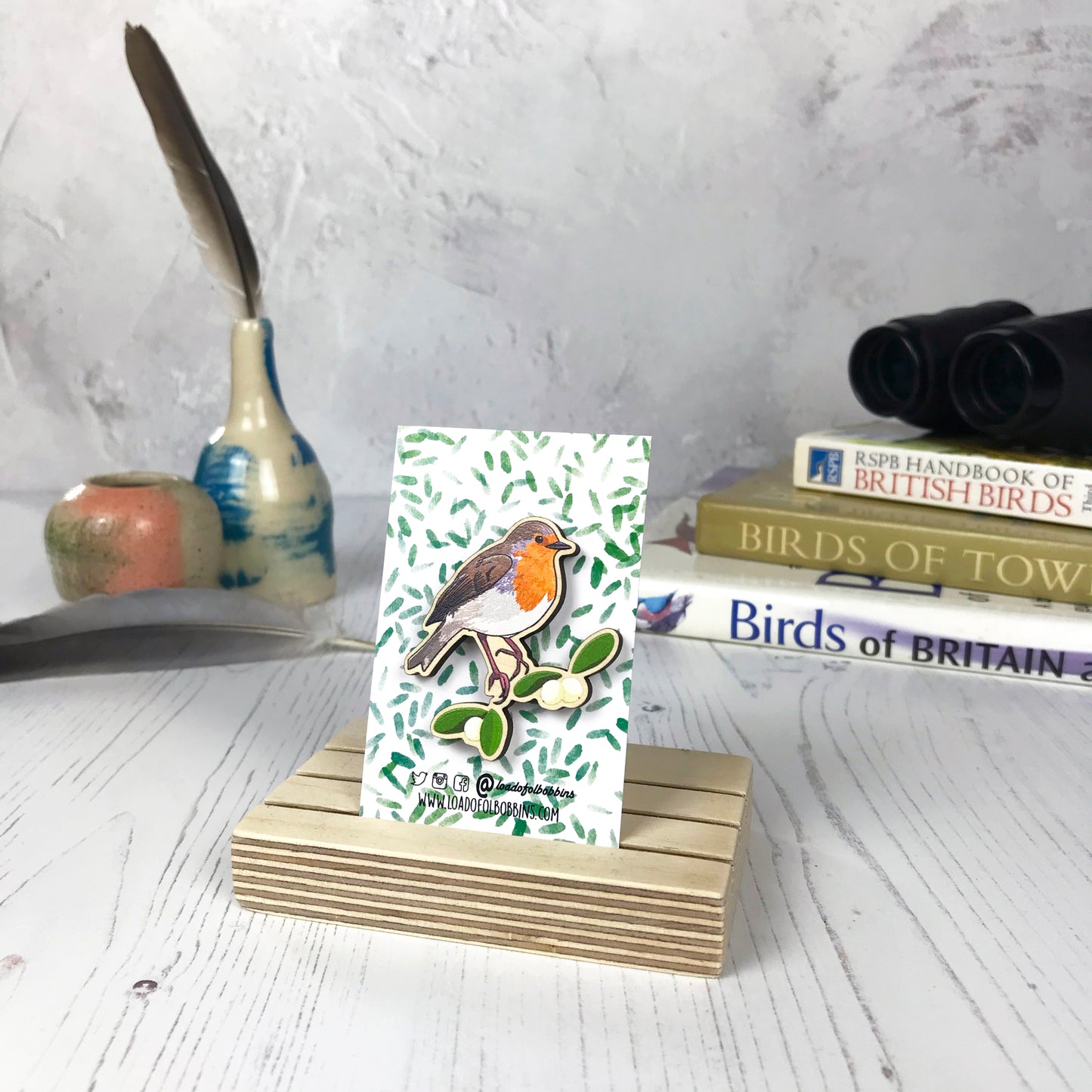 A laser cut wooden brooch showing an illustration of a robin bird perched on some mistletoe by loadofolbobbins. The brooch is mounted on a backing card with a repeat abstract watercolour leaf pattern. They are standing in a wooden stand and in the background you can see a pile of bird watching books and a pair of binoculars.