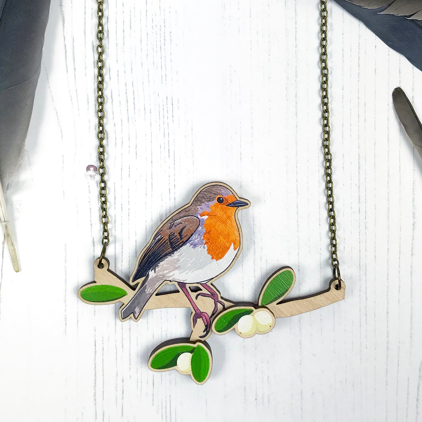A laser cut wooden charm necklace showing an illustration of a robin bird perching on a branch with mistletoe by loadofolbobbins. The mistletoe berries and leaves stand proud of the necklace in a 3D relief effect. The wooden necklace is hanging from an antique bronze cable chain.