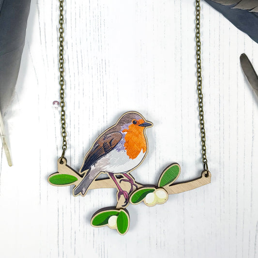 A laser cut wooden charm necklace showing an illustration of a robin bird perching on a branch with mistletoe by loadofolbobbins. The mistletoe berries and leaves stand proud of the necklace in a 3D relief effect.  The wooden necklace is hanging from an antique bronze cable chain.