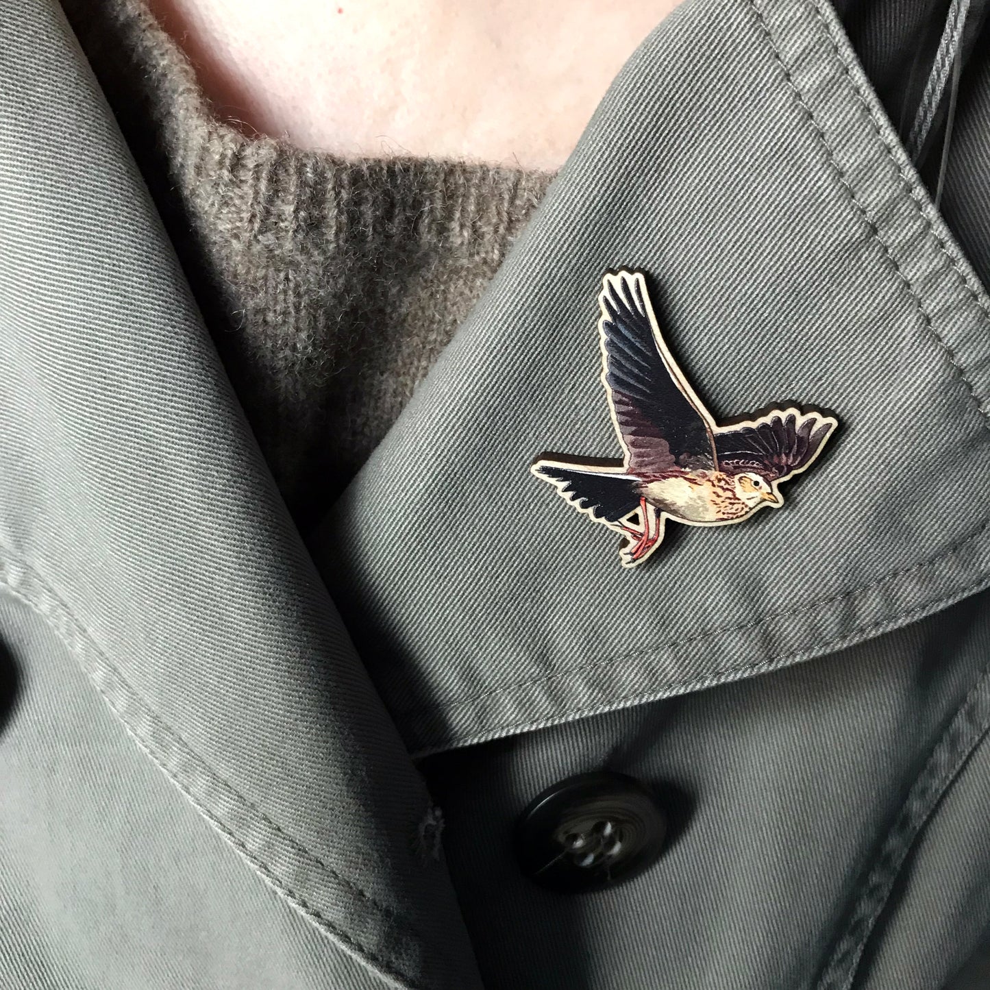 A laser cut wooden brooch showing an illustration by Loadofolbobbins of a flying skylark with wings spread out wide. The brooch is worn on the lapel of a jacket.