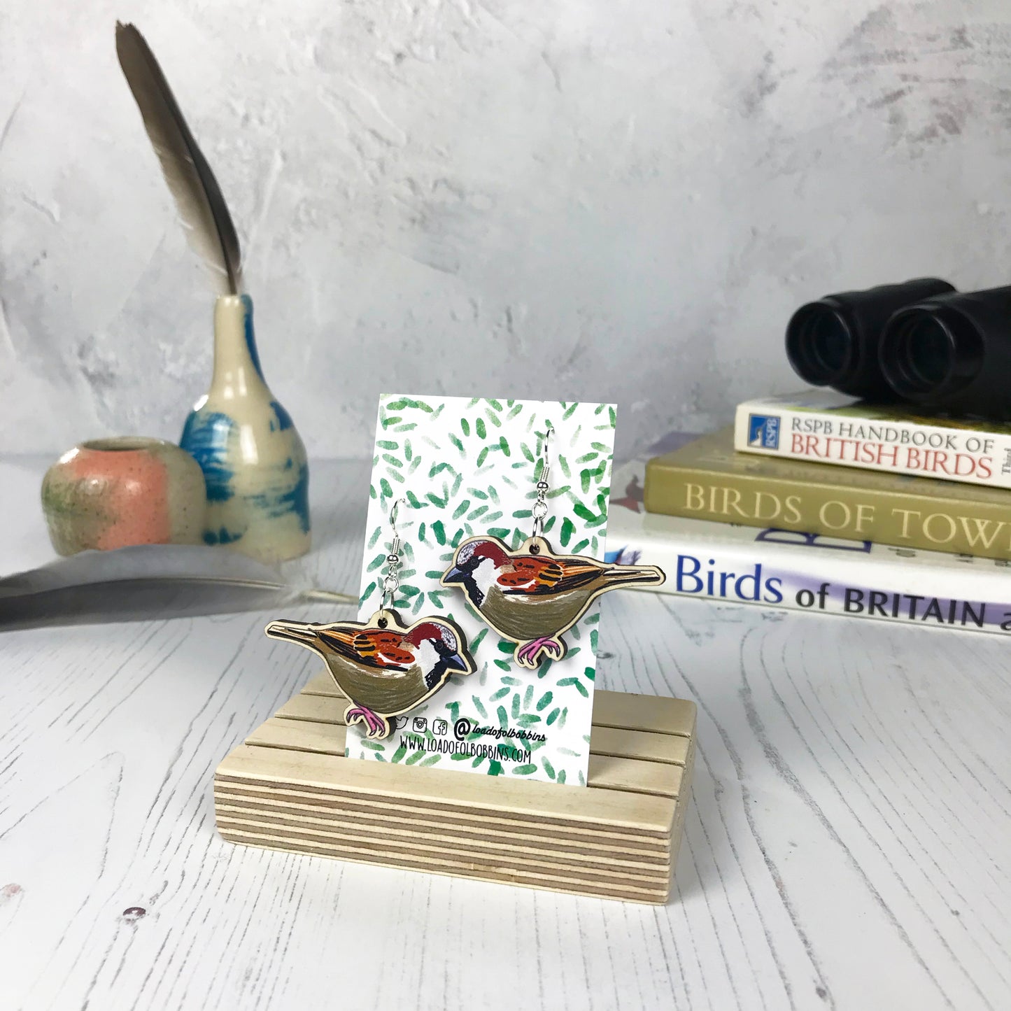 A pair of laser cut wooden dangle earrings showing an illustration of a male sparrow bird by loadofolbobbins. The earrings are mounted on a backing card with a repeat abstract watercolour leaf pattern. They are standing in a wooden stand and in the background you can see a pile of bird watching books and a pair of binoculars.