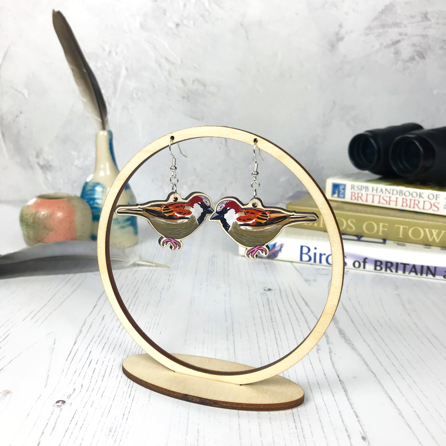 A pair of laser cut wooden dangle earrings showing an illustration of a male sparrow bird by loadofolbobbins. The earrings are hanging from a wooden circular ring display.
