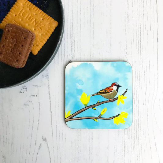 A photo of a round edged square coaster which has an original illustration by Loadofolbobbins showing a male sparrow perched on a forsythia branch with yellow flower blooms. The background is an inky wash in bright turquoise.