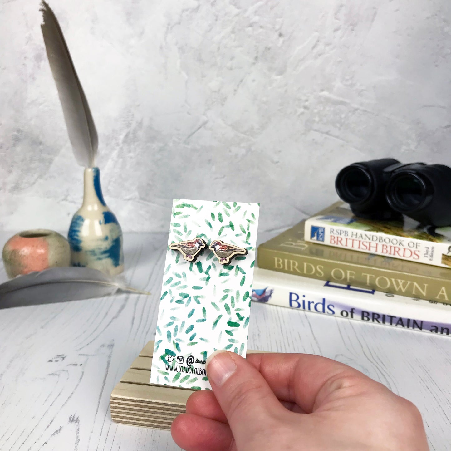 A pair of laser cut wooden earrings showing an illustration of a male sparrow bird by loadofolbobbins. The earrings are mounted on a mini backing card with a repeat abstract watercolour leaf pattern. They are being held by a hand for scale, which shows that each stud earring is slightly smaller than a thumbnail.