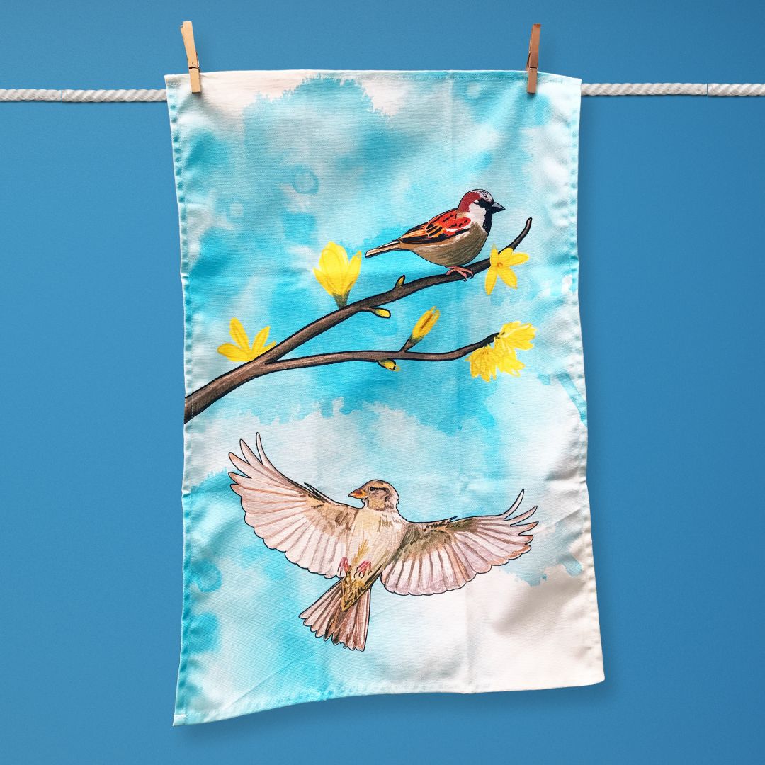 A tea towel hanging on a washing line. The illustration on the tea towel shows a male sparrow perched on a forsythia branch with yellow flower blooms, beneath this is a flying female sparrow with wings spread out. The background is an inky wash in bright turquoise.
