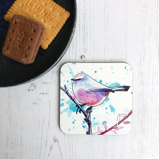 A photo of a round edged square coaster which has an original illustration by Loadofolbobbins showing a white browed tit warbler bird perched on a thin spiky branch. It is quite a puffball shaped bird with an array of pink, blues, purples, oranges and greys in all it’s feathers so it looks like it rolled around in lots of eyeshadows. The background is splashes of light blue ink against white. There are biscuits on a plate in the top left of the photo.